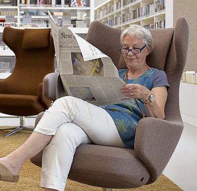 Eine Frau sitzt zurückgelehnt in einem braunen Lesesessel und liest in einer Tageszeitung.