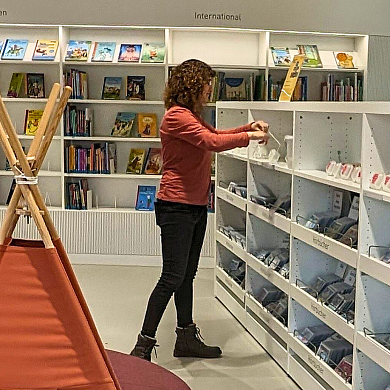 Eine Frau steht inmitten einer Bibliothek und schaut etwas im Regal an.