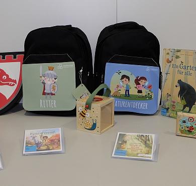 In der Mitte ein schwarzer Rucksack, darum herum sind jeweils ein Buch, eine CD und ein Spielzeug ausgelegt.