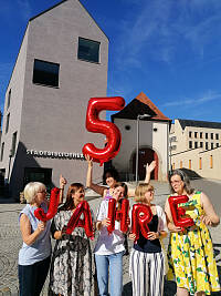 Eine Gruppe von Menschen vor einem Gebäude. Sie halten Luftballons nach oben. Zum einen die Zahl 5 in rot und darunter die Buchstaben JAHRE. 