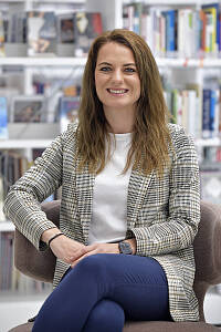 Portrait von einer Frau, sie sitzt in einem Sessel vor einem Bücherregal