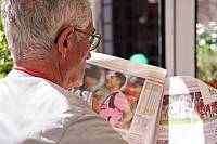 Ein älterer Mann sitzt auf einem Sofa und liest Zeitung. Er ist von hinten zu sehen. 
