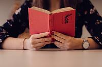 Eine Frau sitzt an einem Tisch und liest ein Buch, das Buch hat einen roten Umschlag.