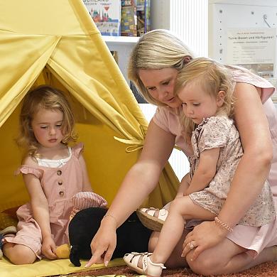 2 kleine Mädchen sitzen gemeinsam mit einer blonden Frau vor einem Kinder-Spielzelt und schauen ein Bilderbuch an